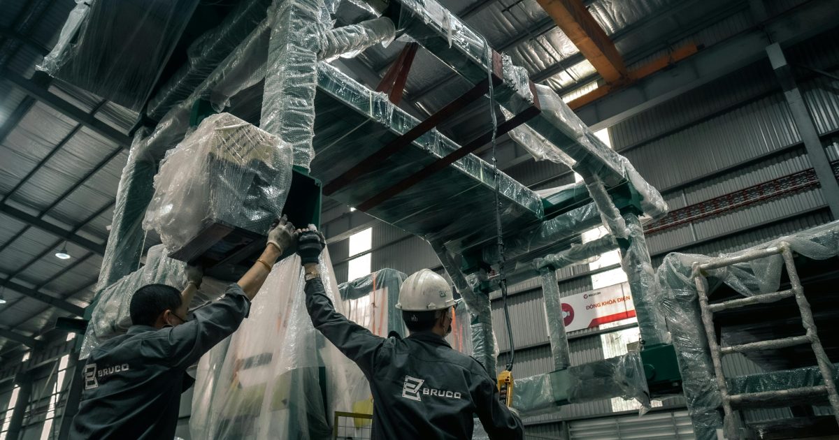 Two workers install large industrial machinery in a warehouse. They wear helmets and work with precision.