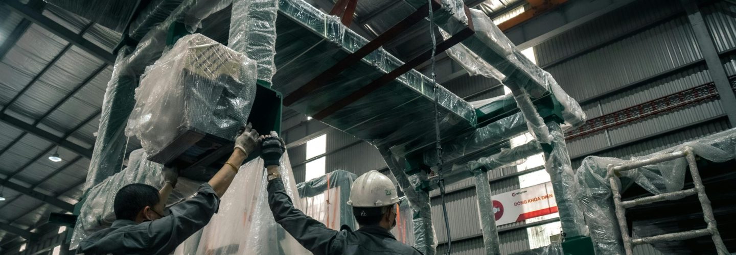 Two workers install large industrial machinery in a warehouse. They wear helmets and work with precision.